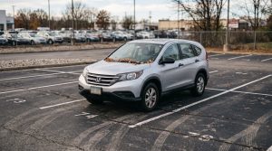 A used Honda CRV parked alone in the back corner of a dealership lot with leaves on the windshield representing aged inventory that has not sold